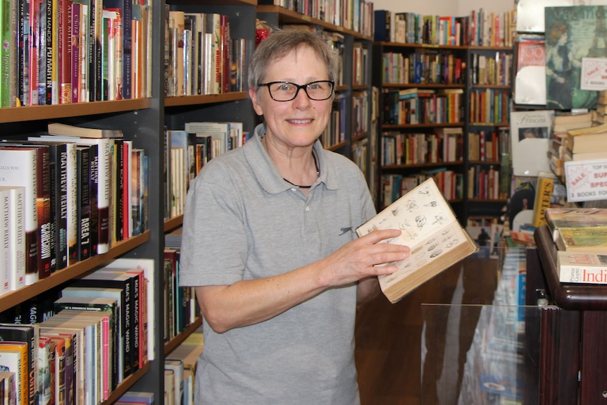 A woman standing among book shelves holds up a book.