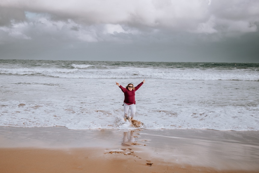 A woman with her hands in the air stands in the water at the beach.