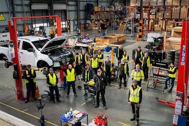 Group of 18 HSV staff in high vis vests stand around white 4x4 ute which is up on hoists with parts off on a factory floor.