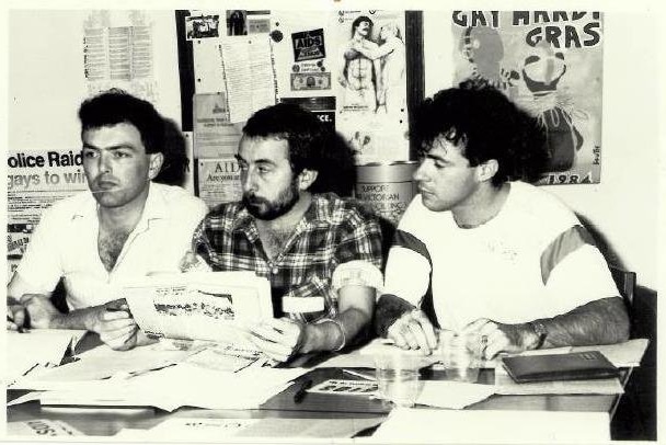 Three young men sit at a desk; the man in the centre holds printed material; posters are pinned behind them.