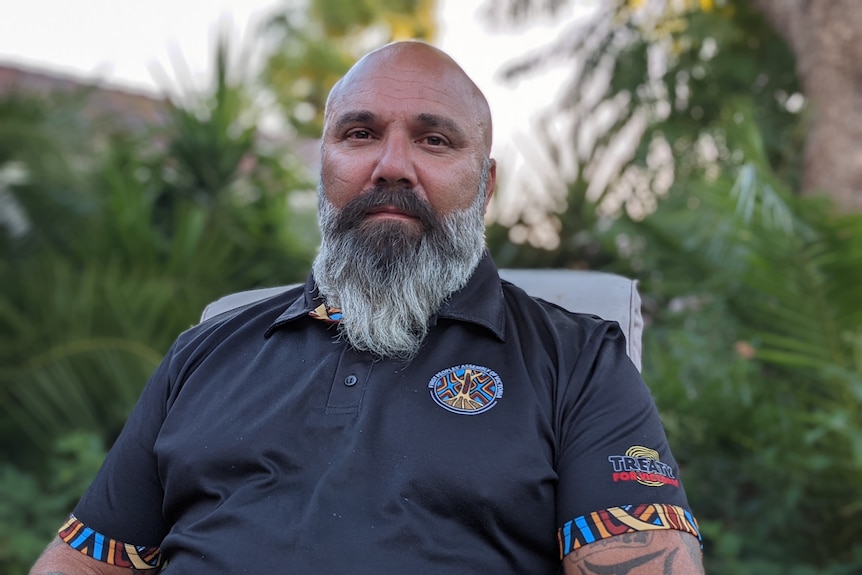 Man with gray beard wearing a black short sleeved collared shirt featuring bands of blue, white, red, orange and yellow stripes.
