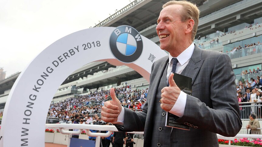 John Size wearing a grey three piece suit and tie with both thumbs up in front of a racecourse grandstand.