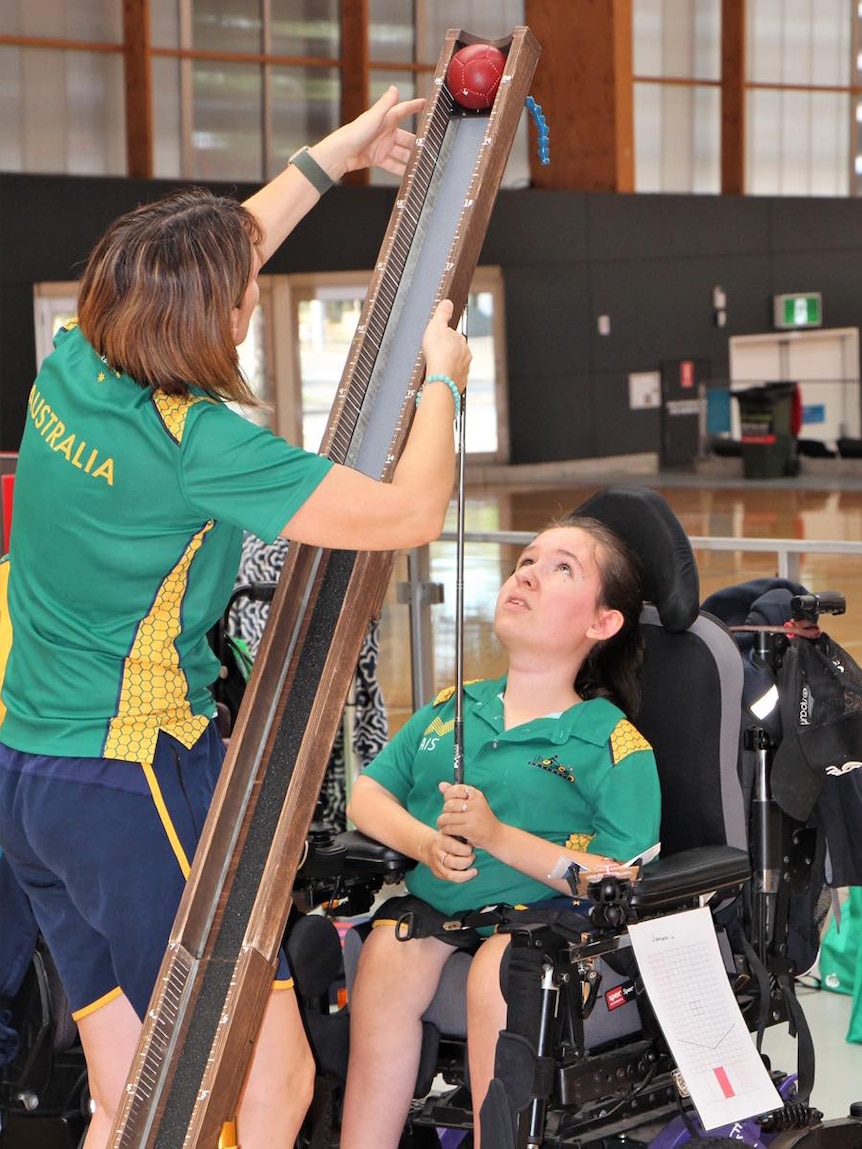 A woman reaches up to hold a round object against a wooden ramp while another woman in a wheelchair looks up at the ramp.