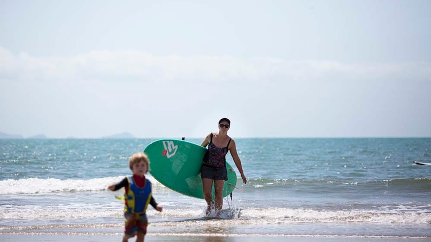 A mother carries a paddleboard out of the surf with her son running ahead of her in the foreground