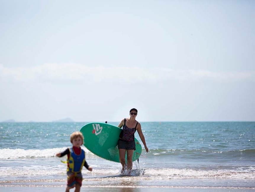 A mother carries a paddleboard out of the surf with her son running ahead of her in the foreground