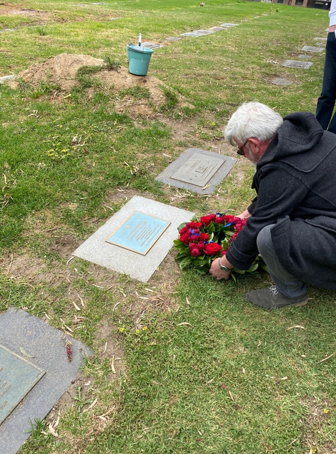 A man squats before the headstone of a grave and places a red-flowered wreath.