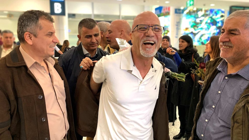 A man wearing a white shirt surrounded by people at an airport arrival gate and smiles broadly as he is helped into a coat.