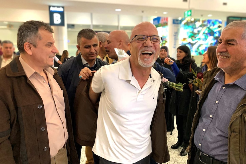A man wearing a white shirt surrounded by people at an airport arrival gate and smiles broadly as he is helped into a coat.