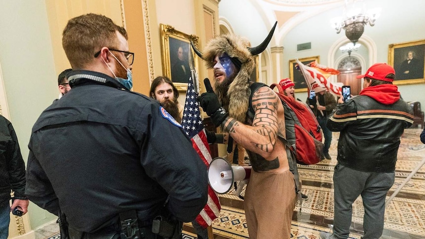 A man wearing an animal hat with horns and the US flag painted on his face confront a police office inside the Capitol.