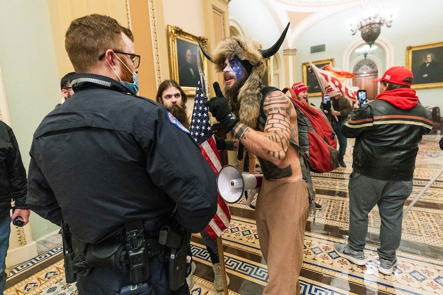 Mężczyzna w zwierzęcym kapeluszu z rogami i amerykańską flagą na twarzy stoi przed biurem policji w Kapitolu.