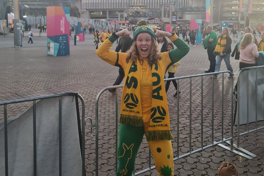 Gemma, who has curly blonde hair, grins, decked out in a Matildas onesie, beanie and scarf.