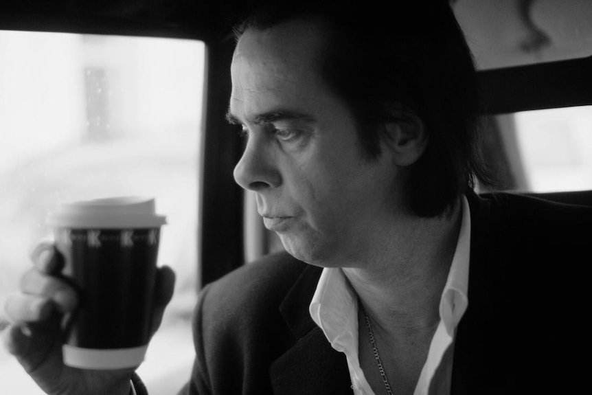 A black and white image of Nick Cave looking sombre, sitting in the backseat of a car, and holding a takeaway coffee