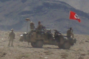 A red swastika flag above a small vehicle with soldiers present and Afghan hills in the background.