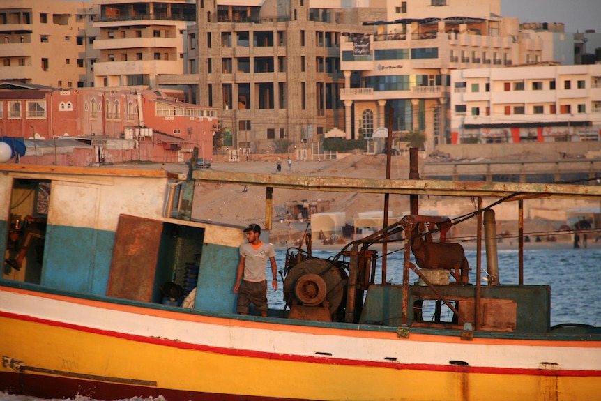 A wide photo of a man standing on an old-looking boat at sea, with some buildings near the shore behind him