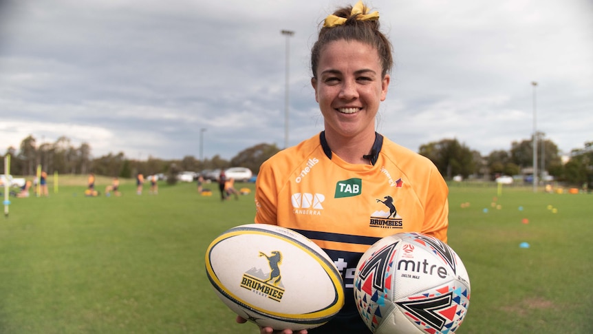 A woman in a rugby jersey holds a soccer ball and rugby ball and smiles at the camera.