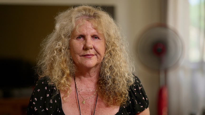 a woman with long curly blonde hair sits looking directly at the camera with a neutral expression