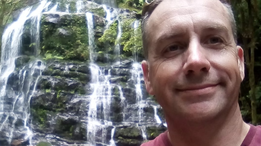 A man takes a selfie in front of a waterfall.