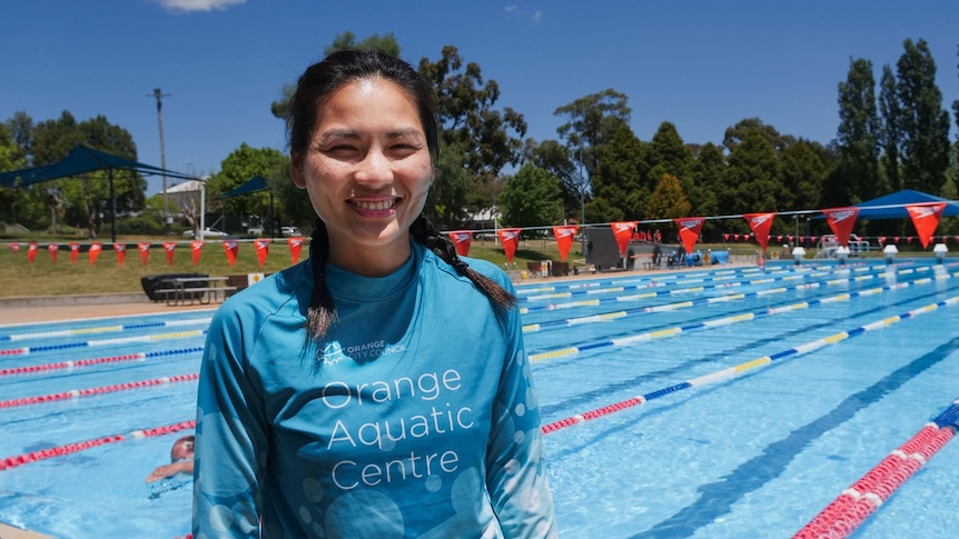 A woman in a blue 'Orange Aquatic Centre' shirt stands in front of an outdoor 50 metre pool.