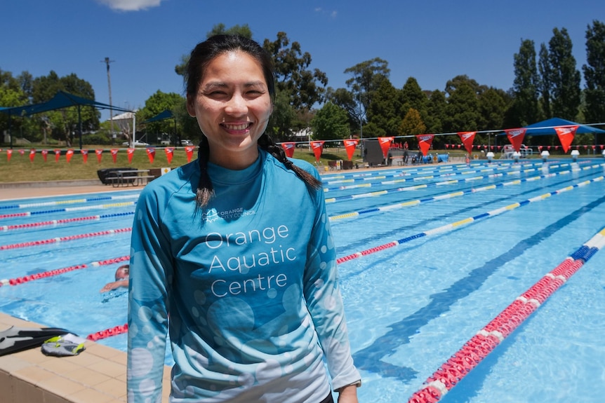 A woman in a blue 'Orange Aquatic Centre' shirt stands in front of an outdoor 50 metre pool.