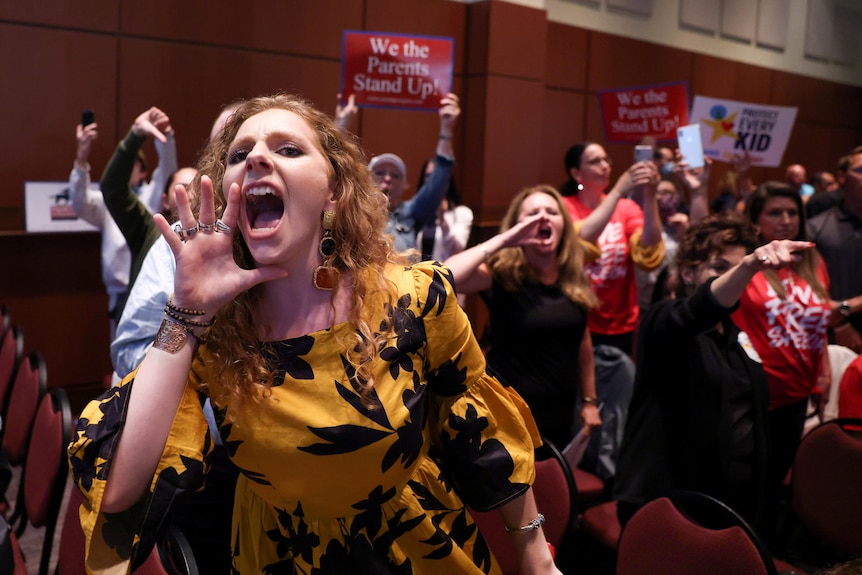 A redheaded woman in a black and yellow printed dress screams in a crowd inside a hall