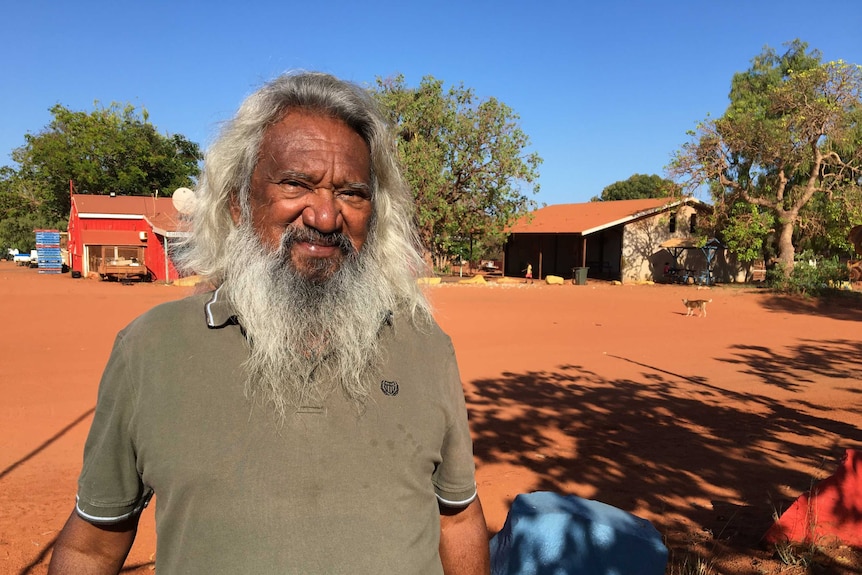 An Indigenous man with long white hair and a long white beard standing outside a desert settlement.