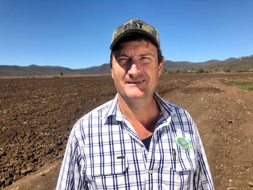 A man wearing a cap and a checked shirt standing in the country on a sunny day.