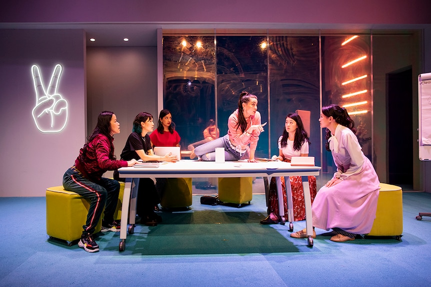 Catherine Văn-Davies sits on table with finger pointed at Mayu Iwasaki as four women look on in office with neon peace sign.