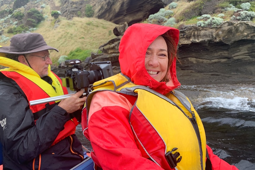 A woman sits in a boat in a life jacket, a cameraman filming passing rocks behind her.