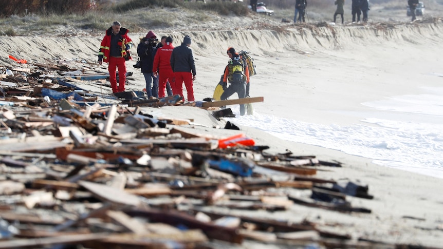 Debris covers sand on a beach as people can be seen gathered in the background. 