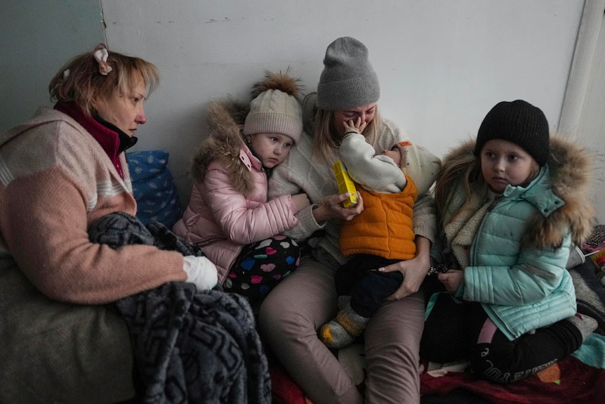 Two women and three young children in winter clothes sit on the floor of a hospital corridor. Some embracing.