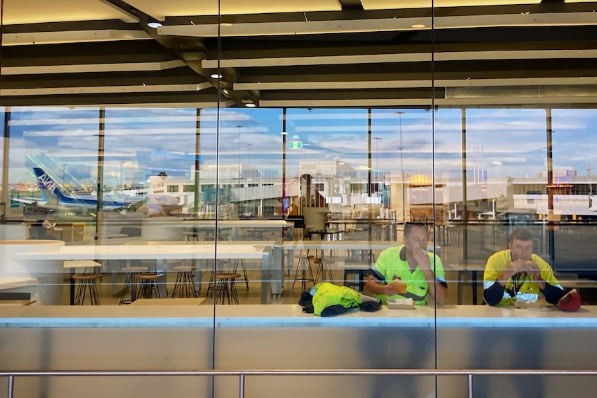 Two men eat a meal at an airport restaurant with a plane reflected in the glass window.