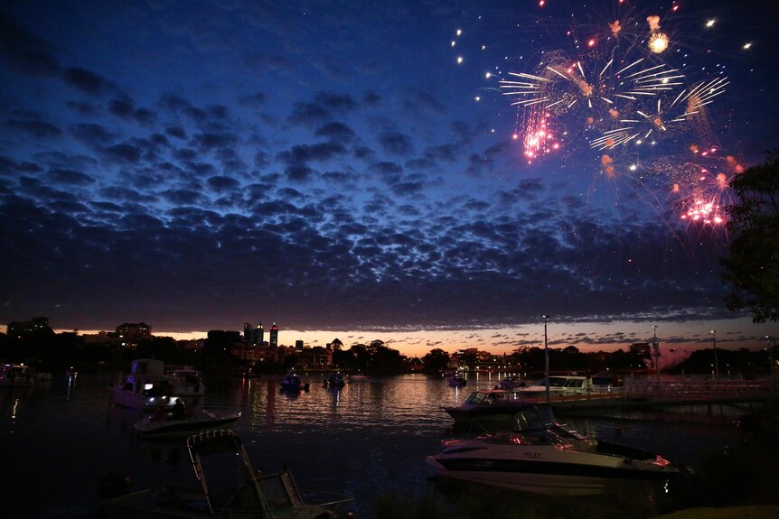 A late sunset photo of fire works exploding in a cloudy sky over the Swan River above several boats before the Perth CBD skyline