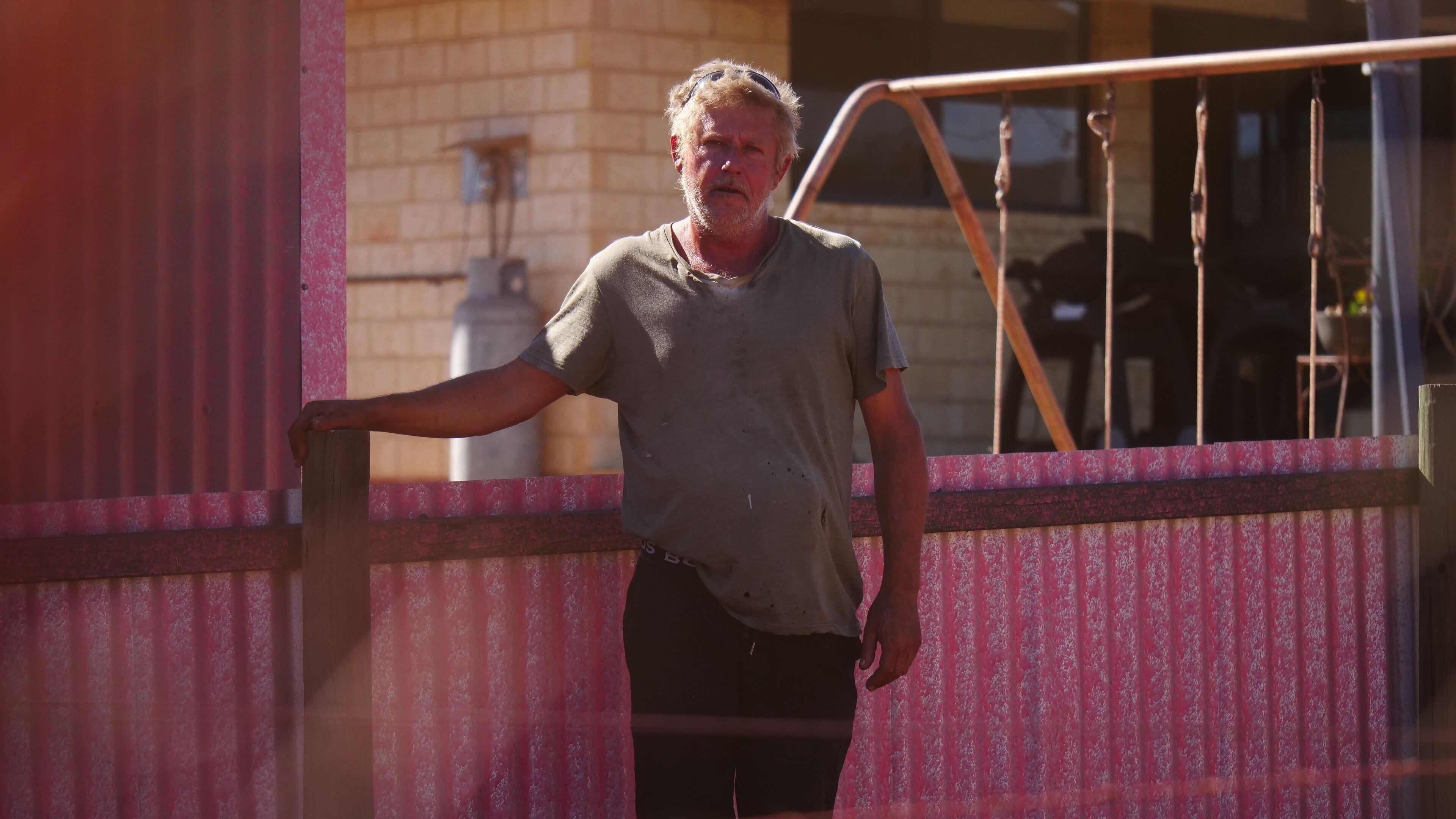 Man in tshirt stands in front of pink-stained corrugated iron fence 
