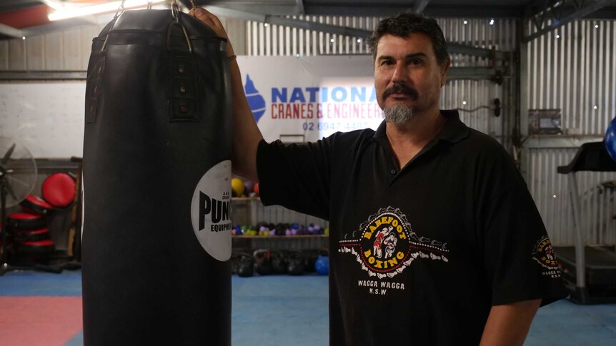 A boxing coach stands, leaning against a punching bag.