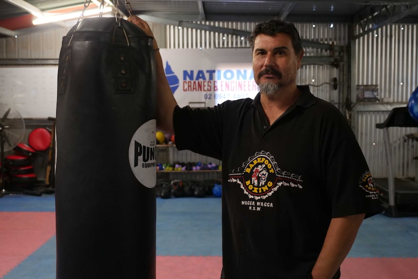 A boxing coach stands, leaning against a punching bag.