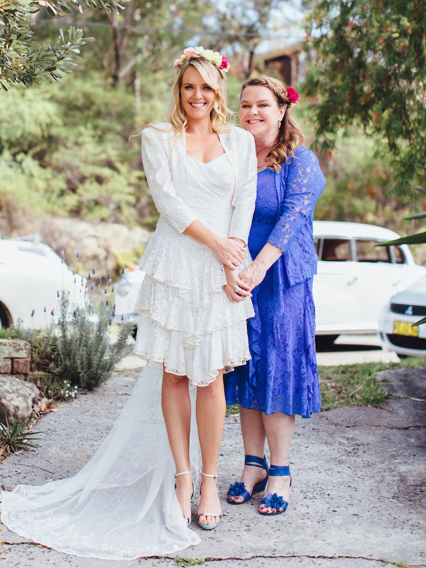 A bride in a white dress with a garland in her hair poses with her mum, in a blue dress, in a bush garden.
