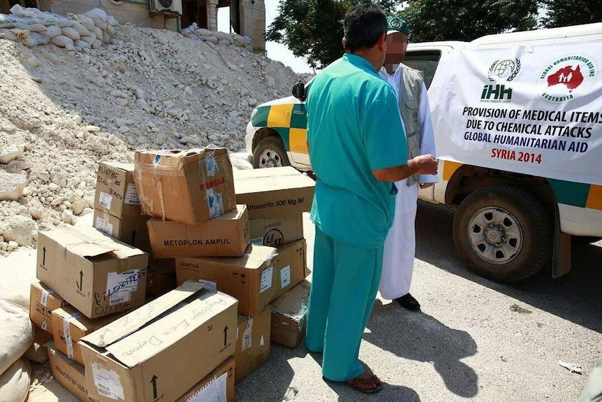 Two men stand next to pile of boxes and small car with banner draped over the side, with rock and rubble in the background