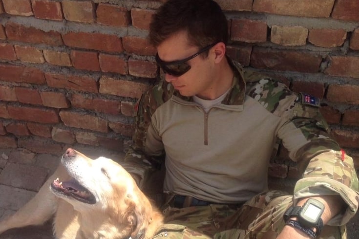 Soldier David Hill sits against a red brick wall and pats a white dog.