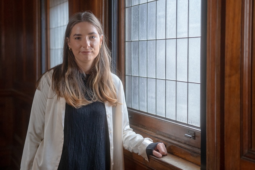 Evelyn Araluen stands next to window with her arm on windowsill, with a slight smile, wearing black blouse and white overshirt