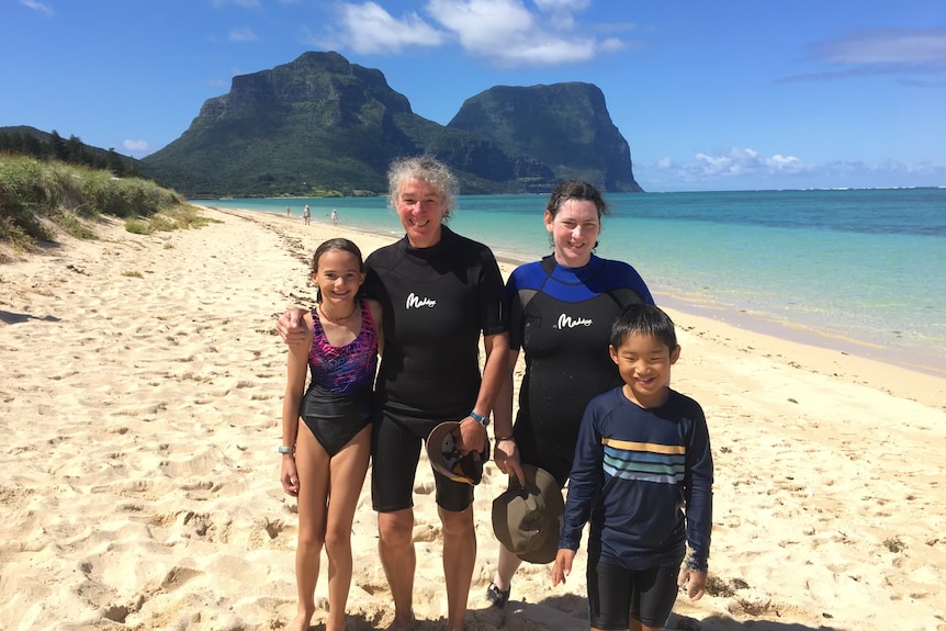 Two swim instructors stand with young children on a beach on an island with a mountainous backdrop.
