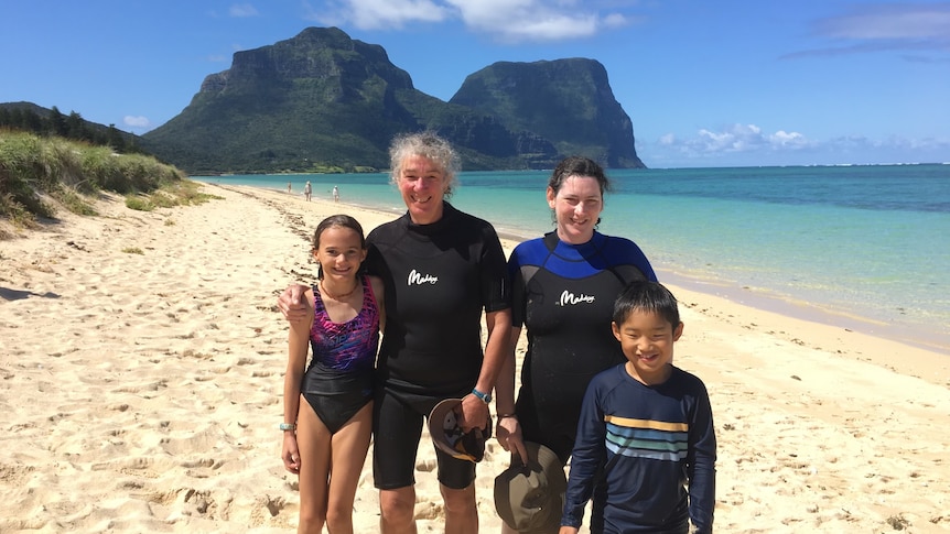 Two swim instructors stand with young children on a beach on an island with a mountainous backdrop.