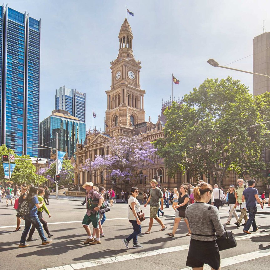 People cross the busy intersection at Sydney Town Hall on a bright, sunny day.