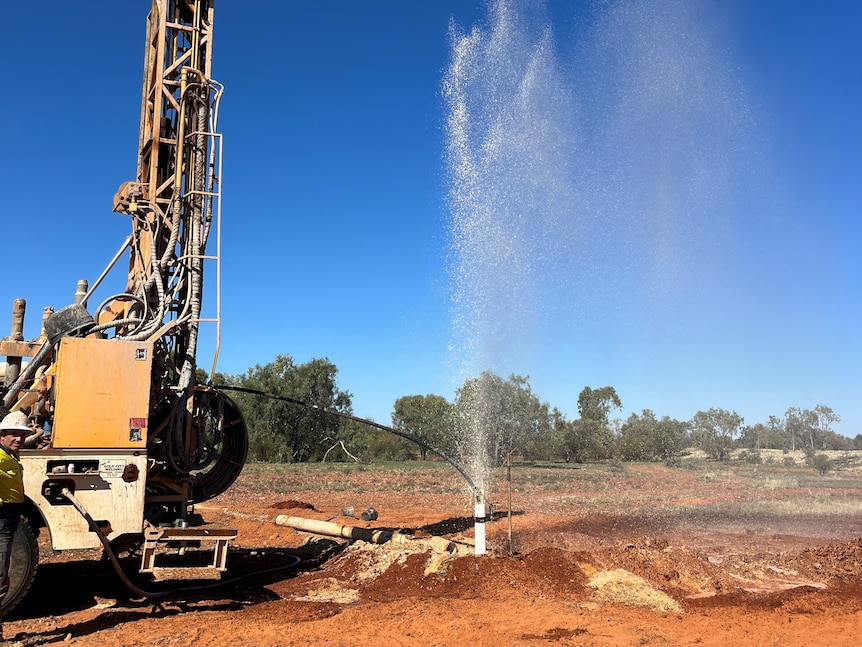 A drill rig stands beside a tall spurt of water coming from the ground after a new bore has been drilled
