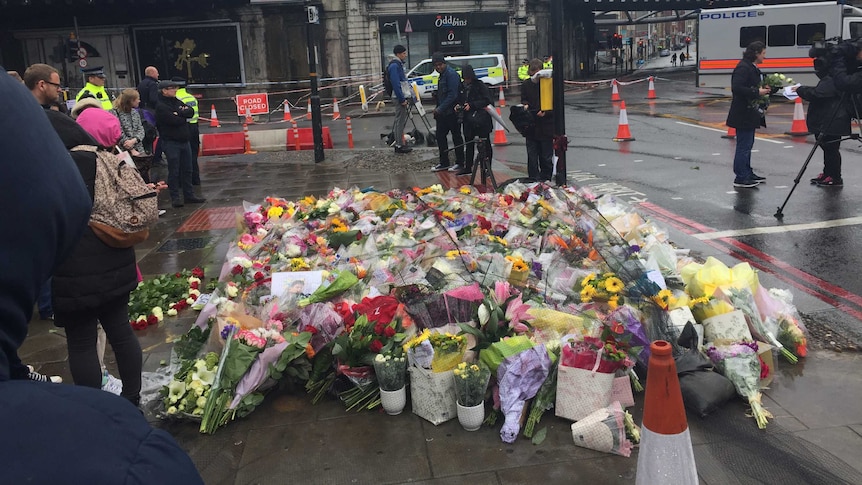 A large collection of flowers sit by the side of the road on a rainy day in London.