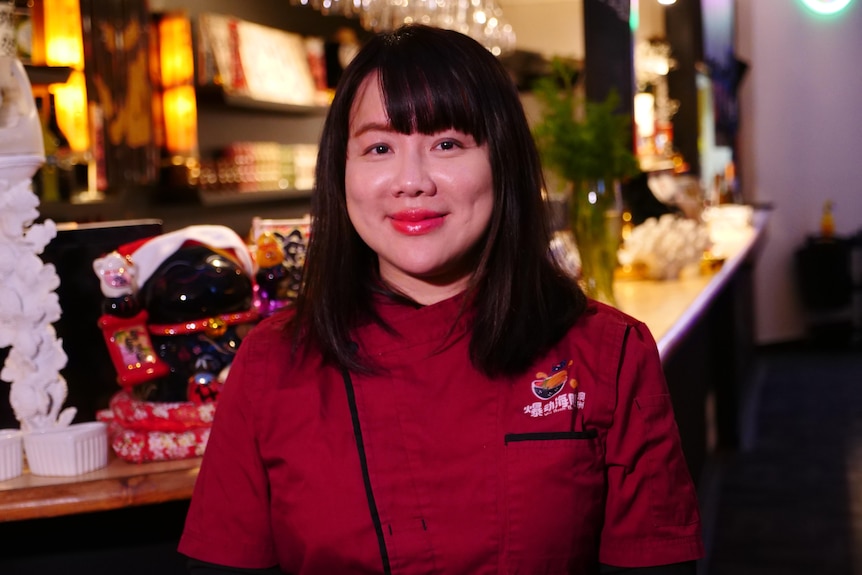 A woman wears a red waiter's outfit and smiles at the camera in a restaurant.