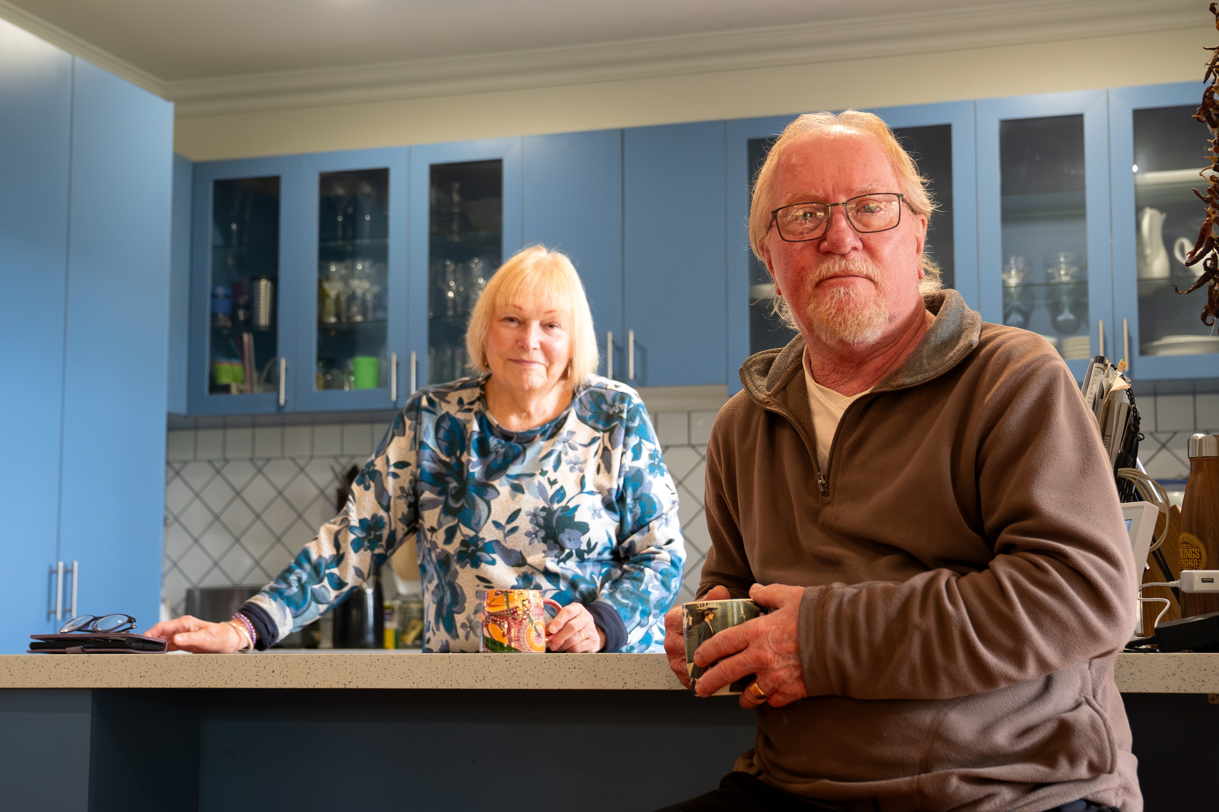 A man and a woman, both with white hair, standing in a blue and white kitchen looking at the camera.