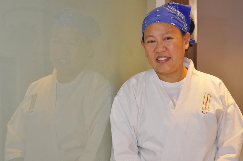 Smiling chef Anita Lo leans against a wall wearing her monogrammed chef's jacket and a bandanna over her hair.