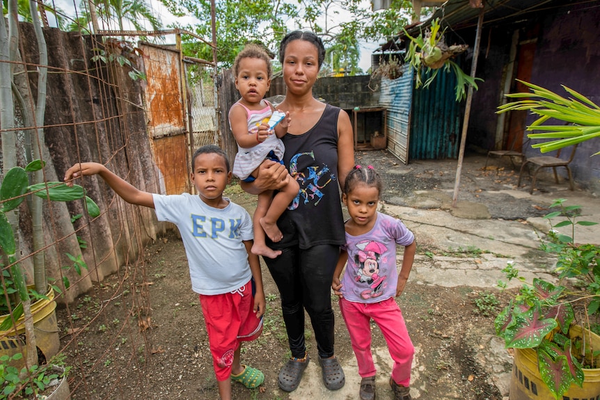 A woman holds a baby, while a young girl and a young boy stand next to her