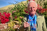 An elderly man in a green knit sweater stands proudly in front of a bougainvillea tree with multiple medals around his neck.