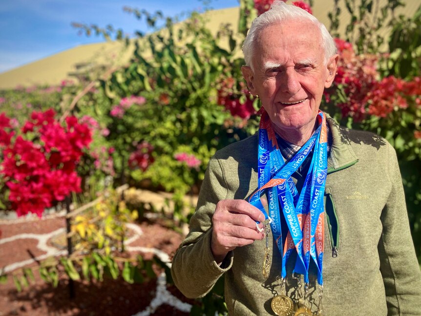 An elderly man in a green knit sweater stands proudly in front of a bougainvillea tree with multiple medals around his neck.